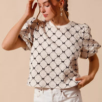 Ivory woven bubble sleeve top with black bow texture pattern, hands-to-ponytail pose