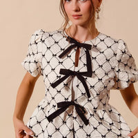 Ivory woven peplum top with black ribbon bows and puff sleeves, close front view.