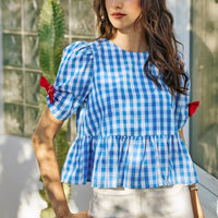 Blue gingham peplum blouse with short sleeves, gathered waist, and red bow ties on shoulders