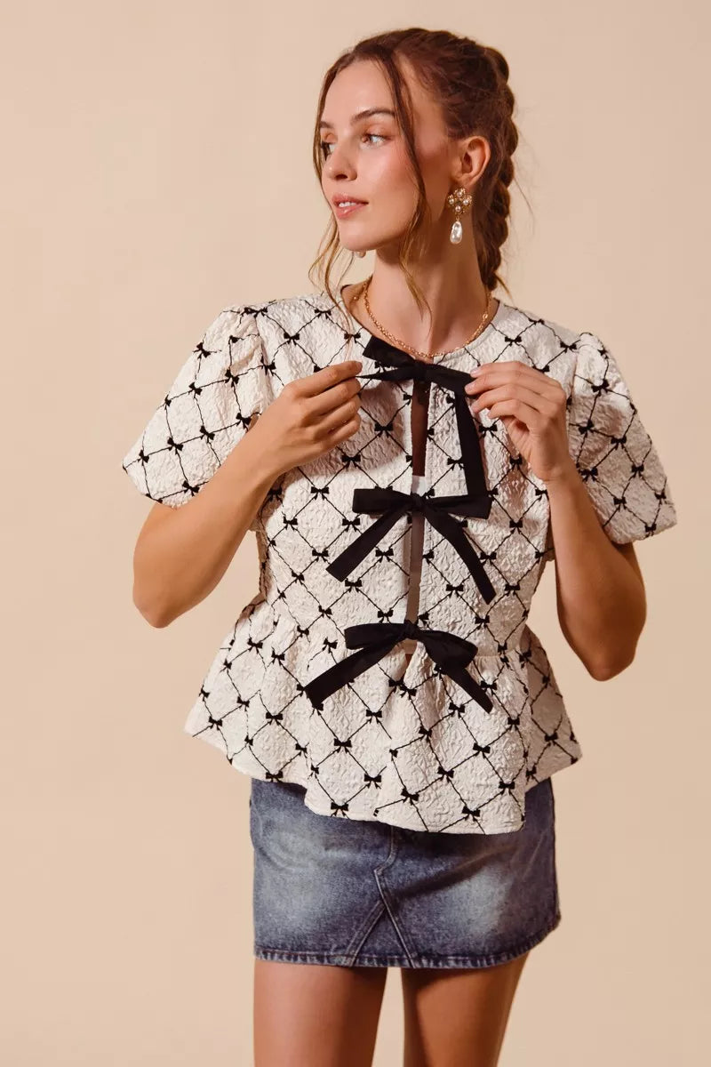 Ivory woven peplum top with black neck bow ties and puff sleeves, front view.