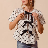 Ivory woven peplum top with black neck bow ties and puff sleeves, front view.