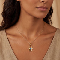 Gold hamsa hand pendant necklace with blue opal-style center, worn at neckline