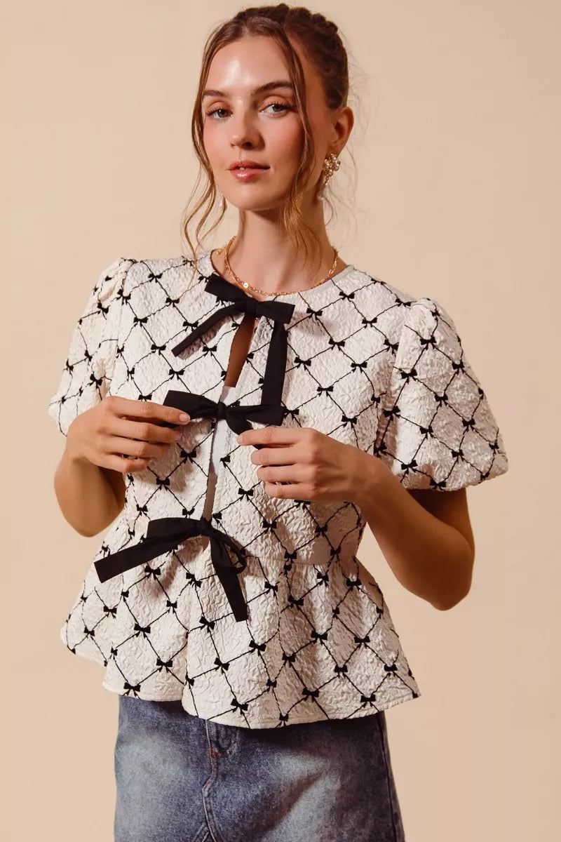 Ivory woven peplum top with black bow tie, puff sleeves, and lattice ribbon print.