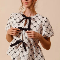 Ivory woven peplum top with black bow tie, puff sleeves, and lattice ribbon print.
