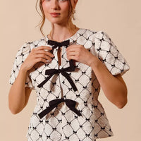 Ivory woven peplum top with black bow tie at neckline and puff sleeves, front view.
