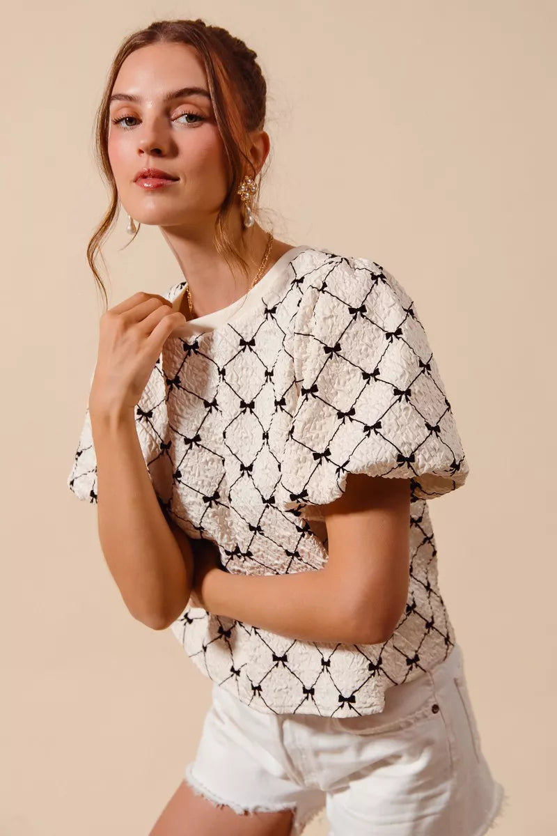 Ivory woven bubble sleeve top with black bow texture pattern and cropped shoulder detail