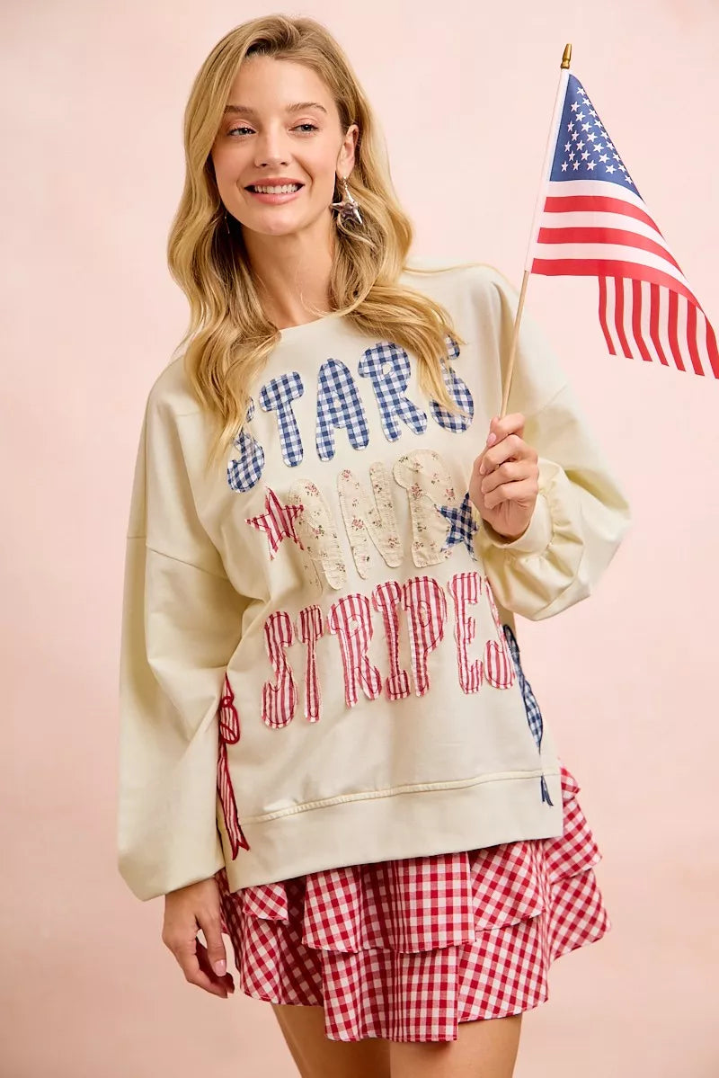 Cream sweatshirt with star-and-stripe patch lettering and red-blue bow graphic on side.