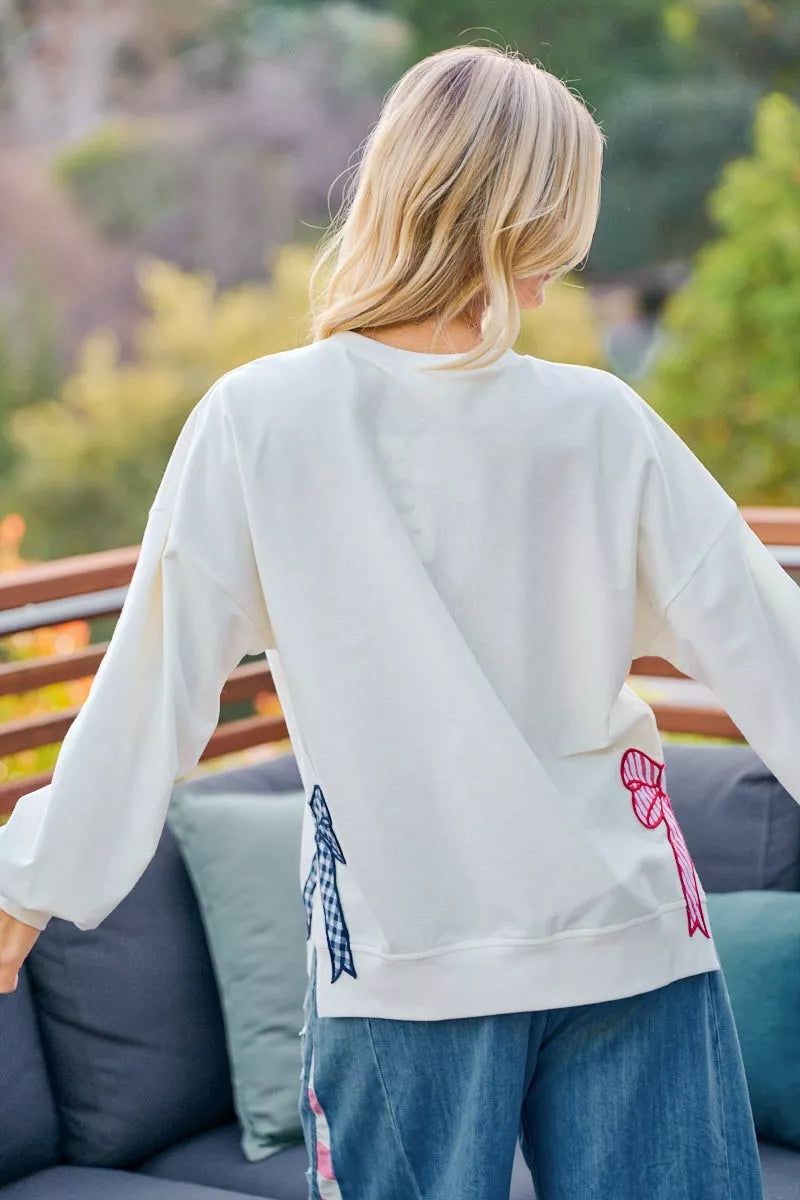 Cream sweatshirt back showing red and blue bow patches and side seam stitching.