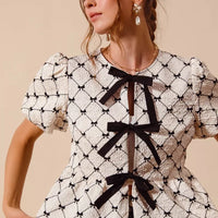 Ivory woven peplum top with black bow tie ribbons and short puff sleeves, angled view.