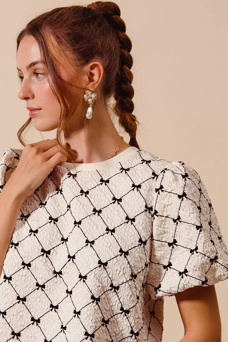 Ivory woven bubble sleeve top with black bow texture pattern, sleeve and neckline close-up