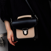 Black leather crossbody handbag with beige flap and silver buckle, worn by hand