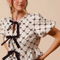 Ivory woven peplum top close-up with black bow ties at neckline and lattice print.