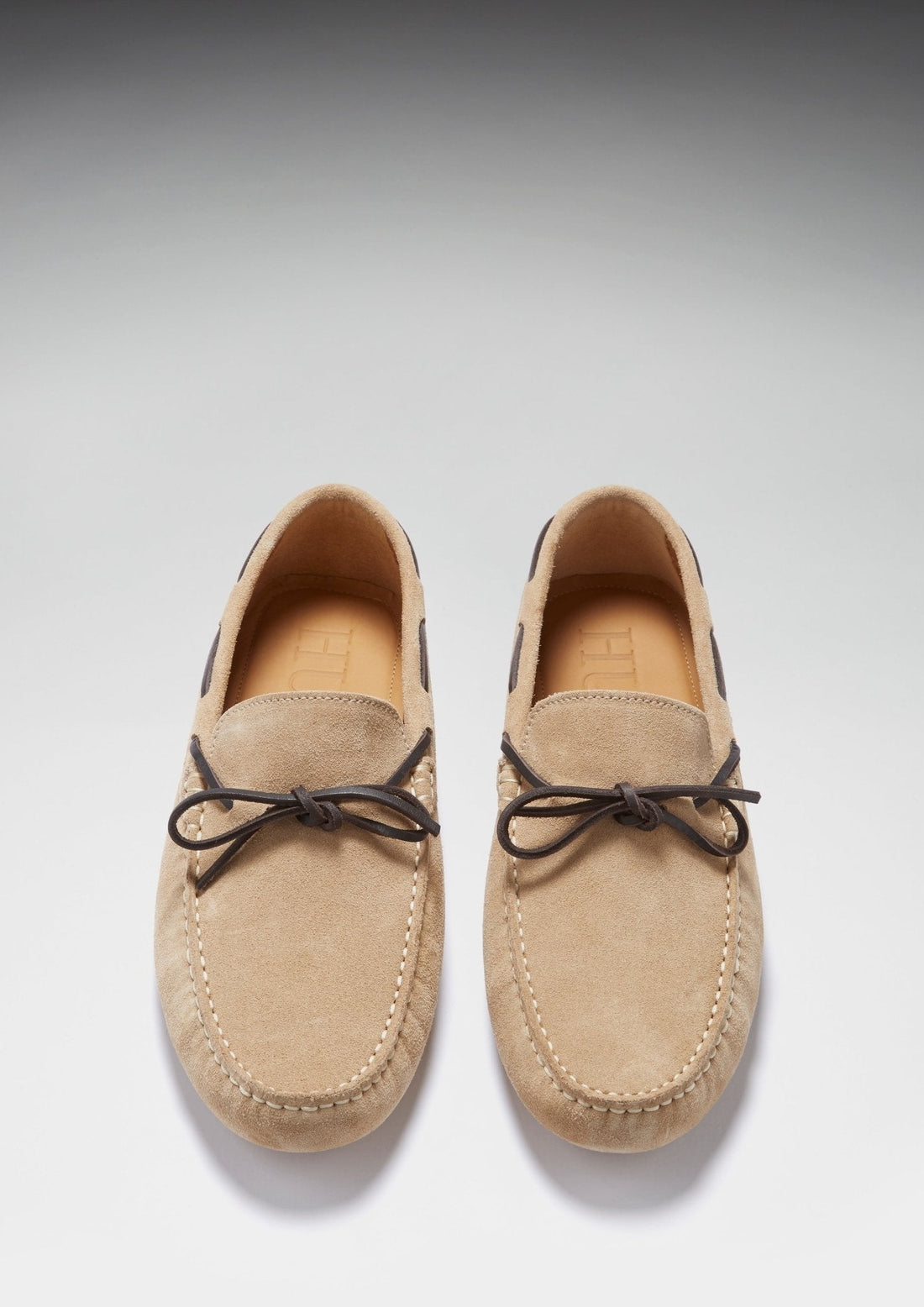 Pair of taupe suede driving loafers viewed from above, with dark bow laces and stitched toes